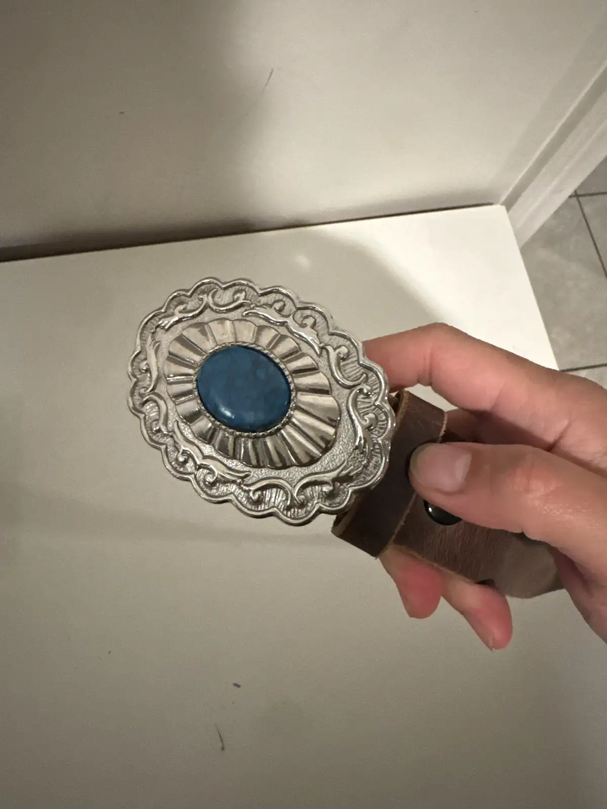 A hand holds up a large, ornate silver belt buckle with a blue stone center, attached to a brown leather strap, against a…