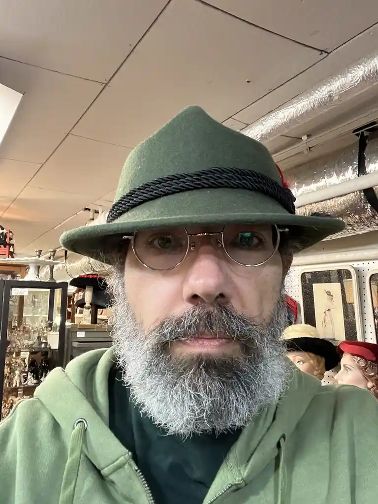 A man with a gray beard and round glasses wears a green felt hat with a black braided band, looking directly at the camera…