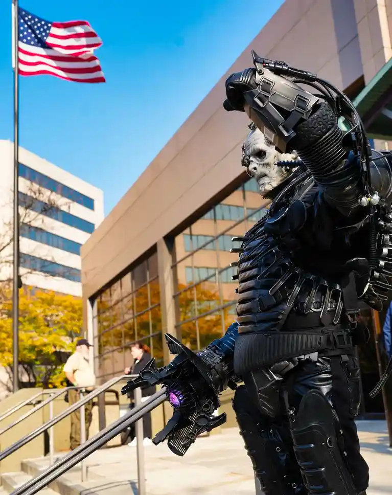 A person in a detailed, black, futuristic armored costume with a skull-like mask and glowing purple gauntlet stands outside…