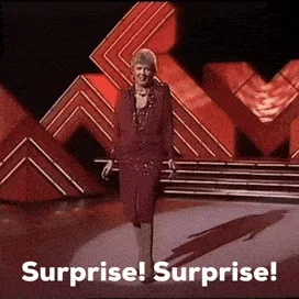 A woman in a dark purple suit walks confidently across a stage with red geometric set pieces behind her. In each of the six…