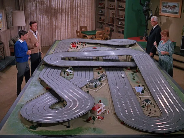 A still from a 1960s TV show shows four people standing around a large, elaborate model slot car racetrack with multiple…