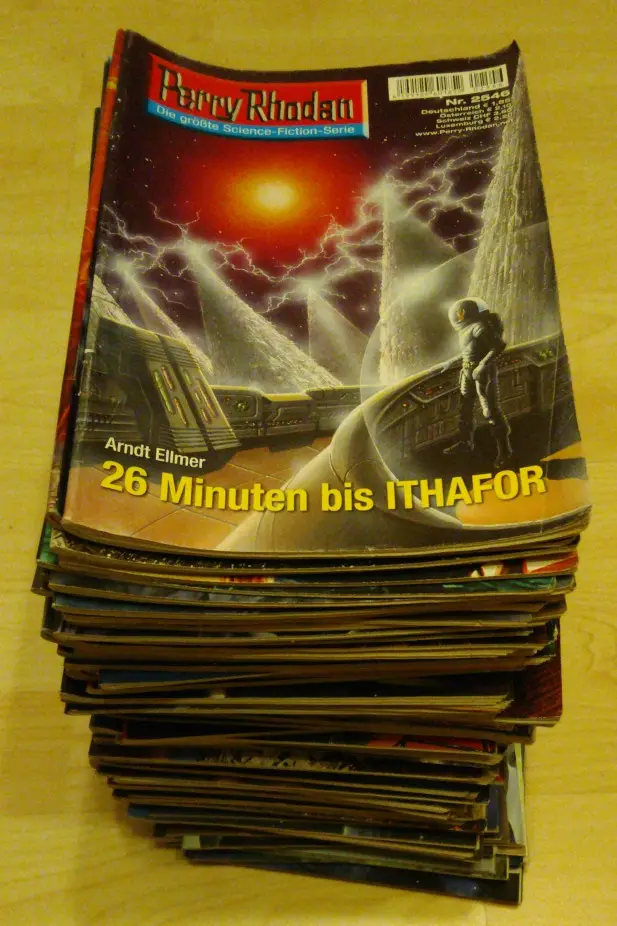 A tall stack of Perry Rhodan science fiction magazines rests on a wooden surface, with the top magazine's cover showing a…