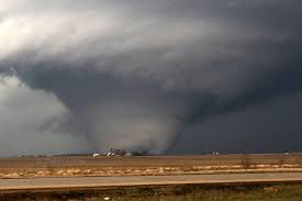 A wide shot of a large, dark tornado funnel cloud touching down in a flat, rural landscape with a road in the foreground…