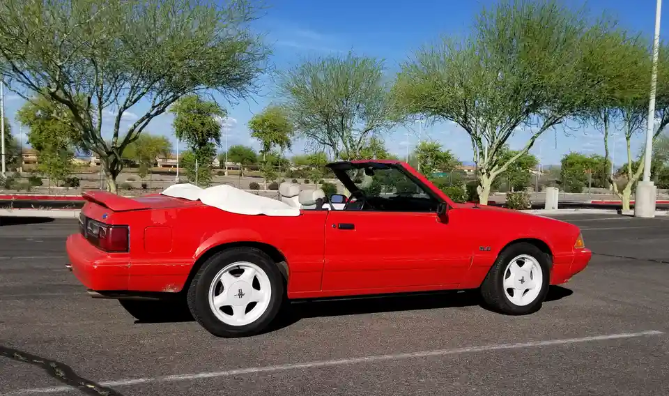 A red 1980s Ford Mustang convertible with a white soft top folded behind the seats is parked in a sunny lot with green…