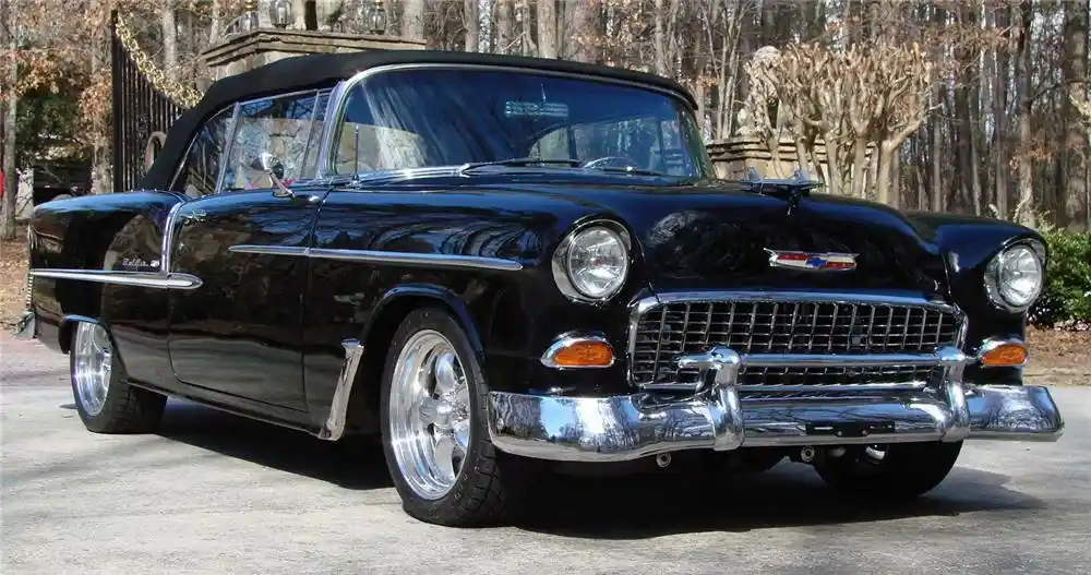 A glossy black 1957 Chevrolet Bel Air convertible with chrome bumpers, whitewall tires, and a black soft top is parked on a…