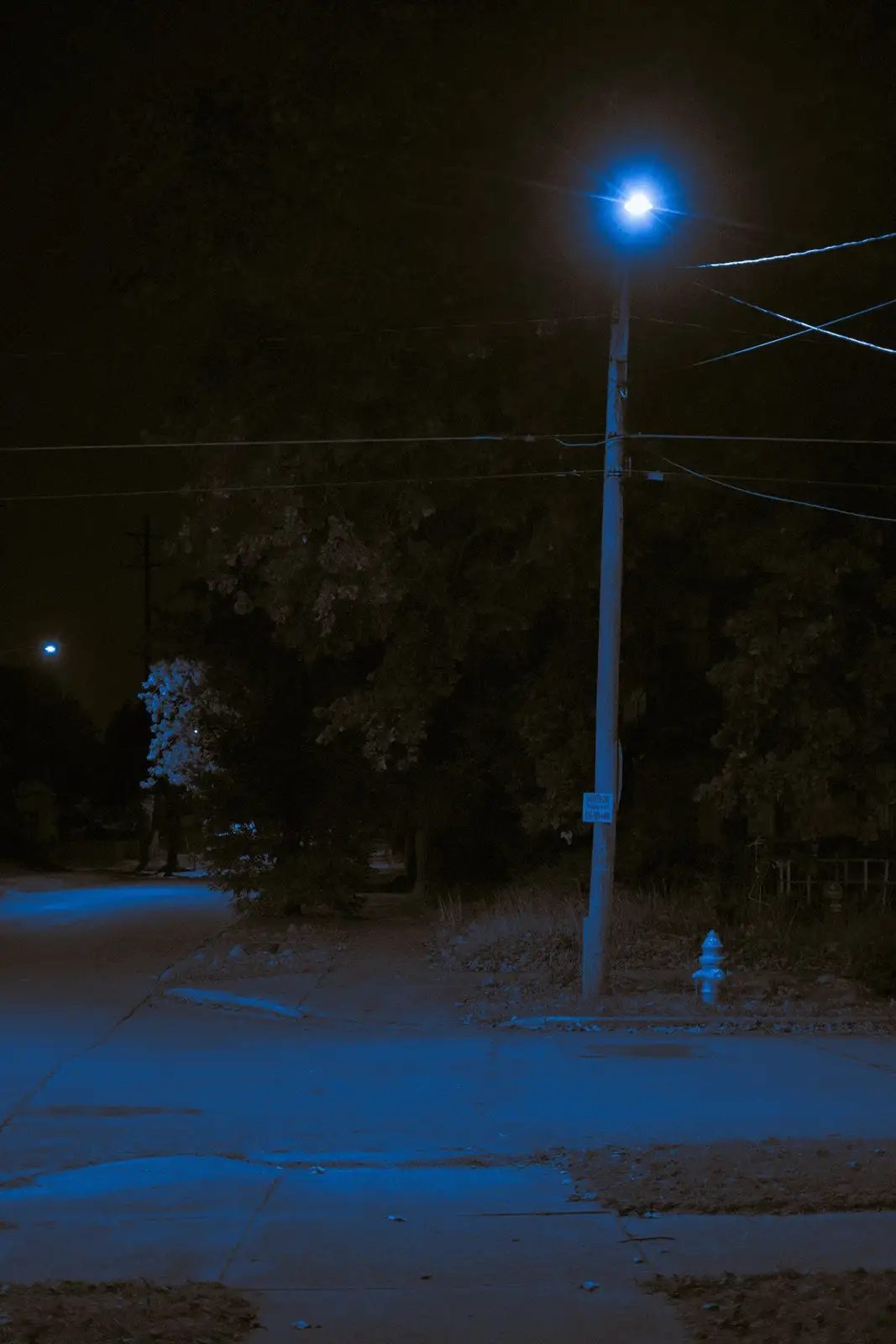 A nighttime street scene dominated by a single, intensely bright blue streetlight casting a cool glow on the pavement…