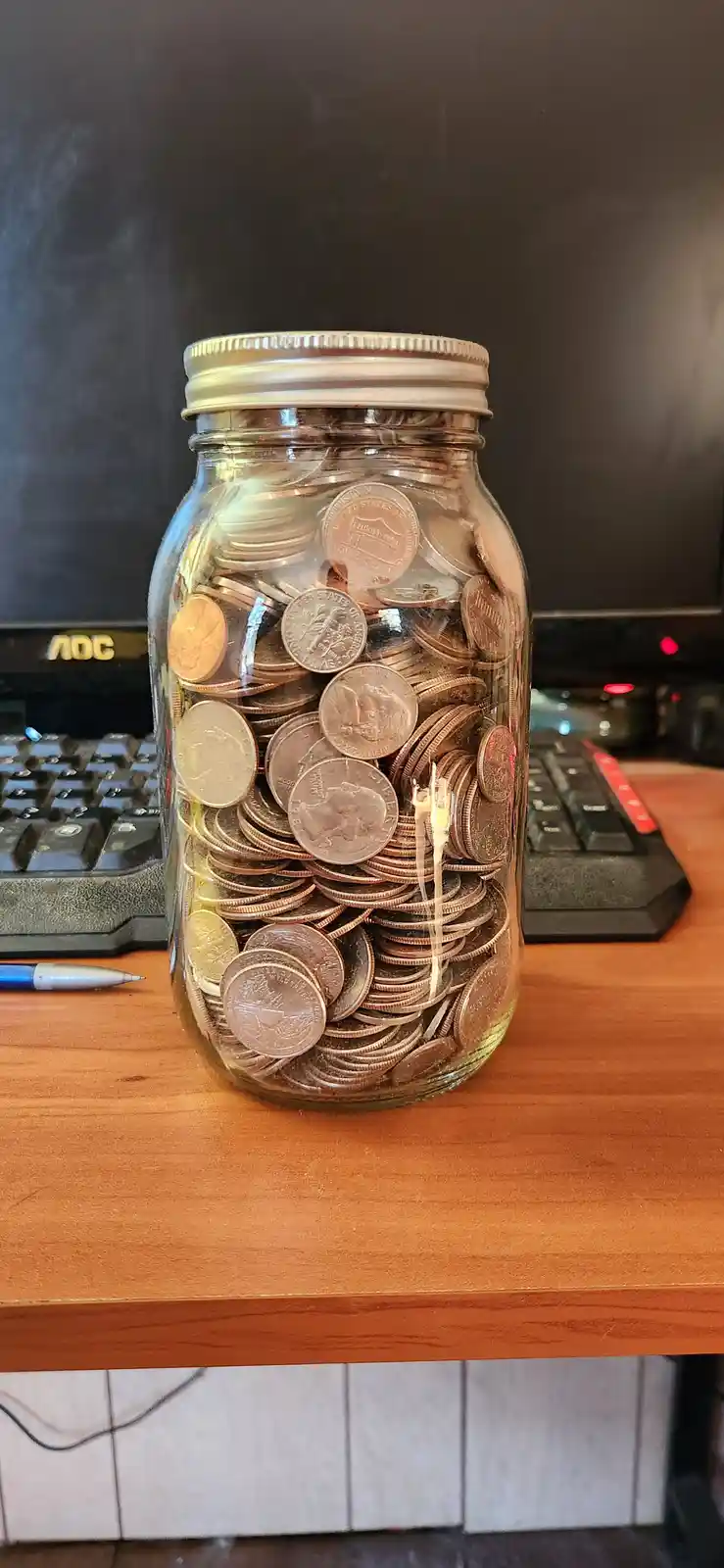 A clear glass mason jar filled to the brim with various U.S. coins sits on a wooden desk, with a computer monitor and…