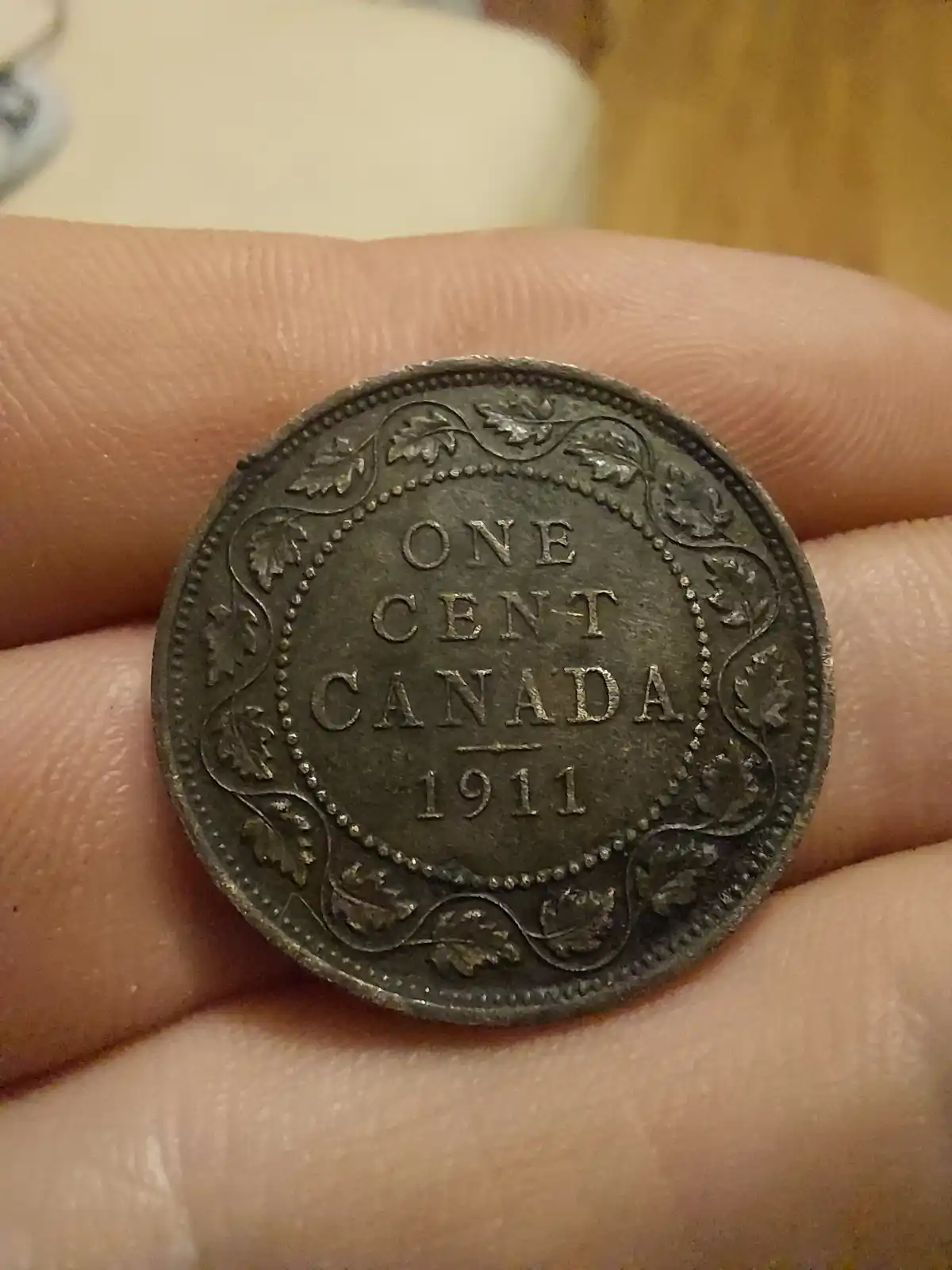 A close-up photo shows a hand holding a tarnished 1911 Canadian one-cent coin, which reads 'ONE CENT CANADA 1911' in raised…