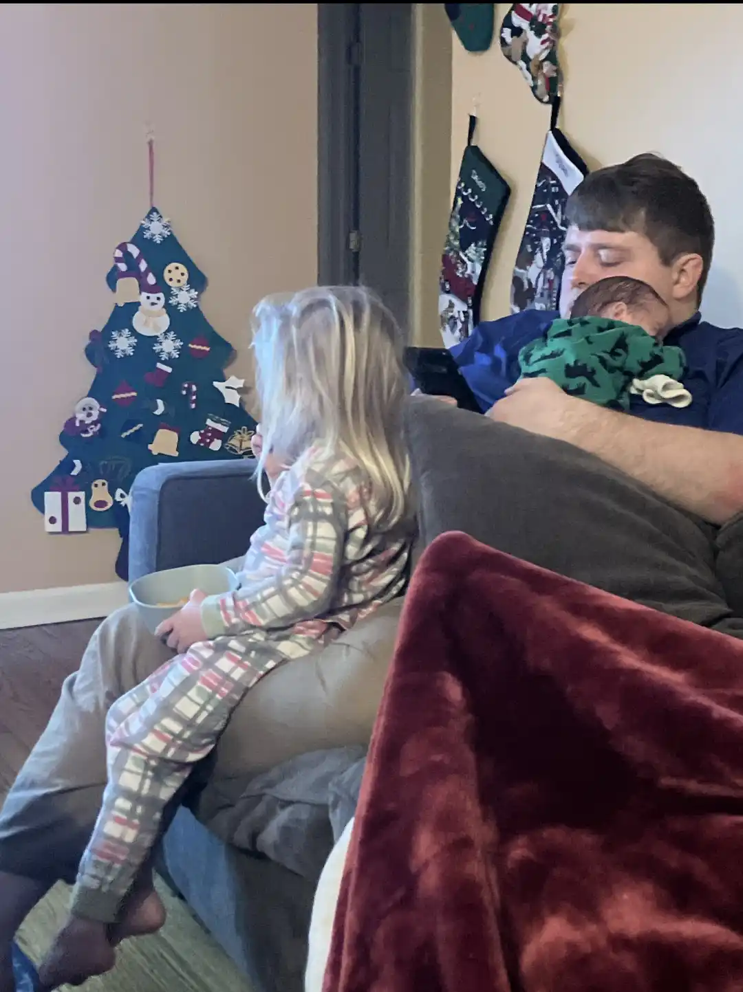 A man in a blue shirt is sitting on a couch holding a sleeping baby wrapped in a green blanket, while a young girl in plaid…