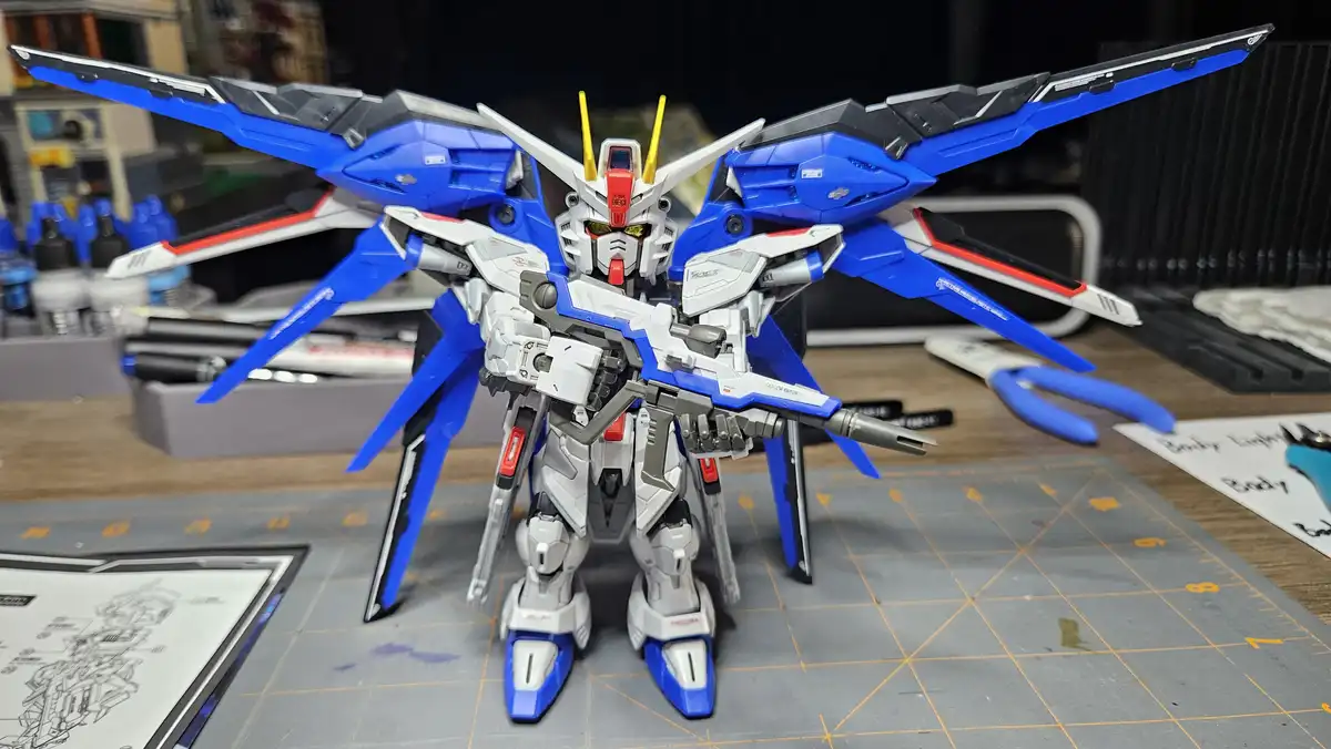 A blue, white, and red Gundam model kit stands on a grey grid mat surrounded by modeling tools, paint bottles, and…