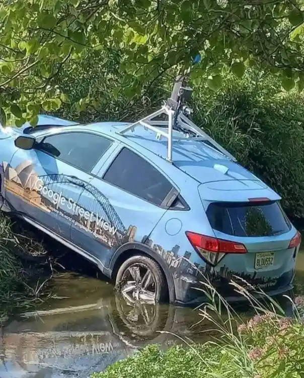 A light blue Google Street View car is stuck at an odd angle in a muddy ditch surrounded by green foliage. The vehicle's…