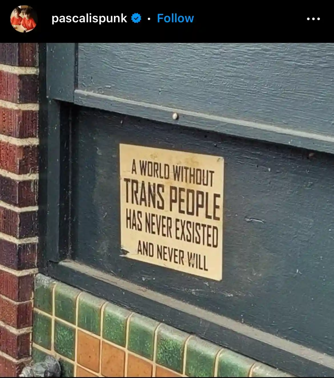 A close-up photo shows a beige sign with bold black text affixed to a dark green wooden door frame next to a brick wall and…