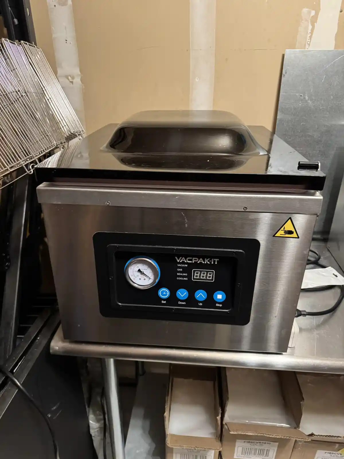 A stainless steel VACPAK-IT vacuum sealer sits on a metal shelf in a commercial kitchen, its digital display showing '8.8.8'…