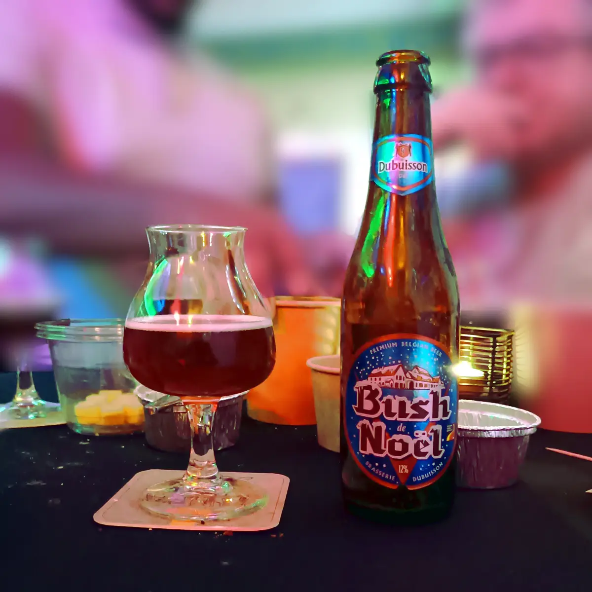 A bottle of Dubuisson Bush de Noël Belgian beer sits next to a glass filled with the dark red beverage on a coaster…
