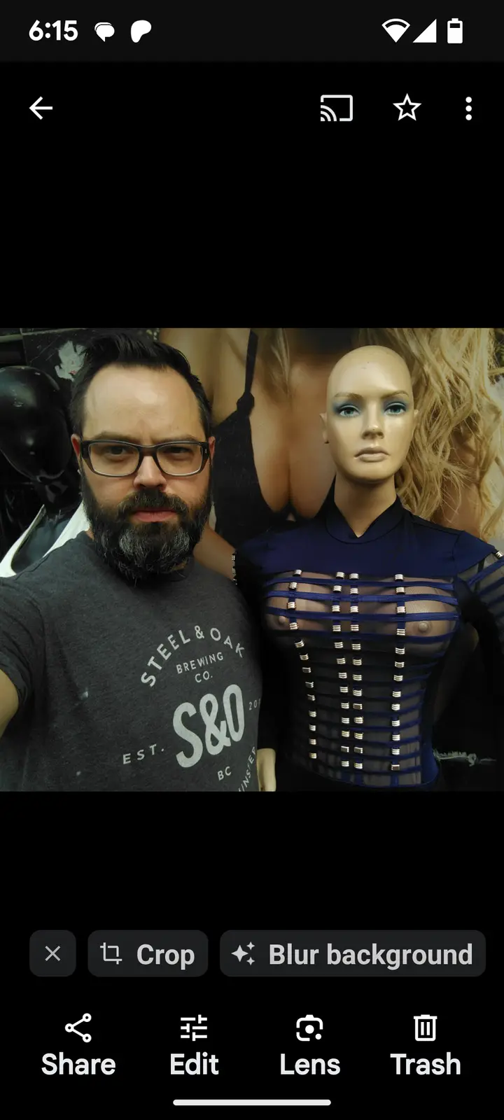 A man with glasses and a beard takes a selfie next to a female mannequin wearing a revealing blue outfit. The man has a…