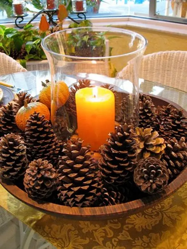a wooden bowl filled with pinecones, small pumpkins, and a lit yellow candle inside a glass hurricane vase, arranged as a…