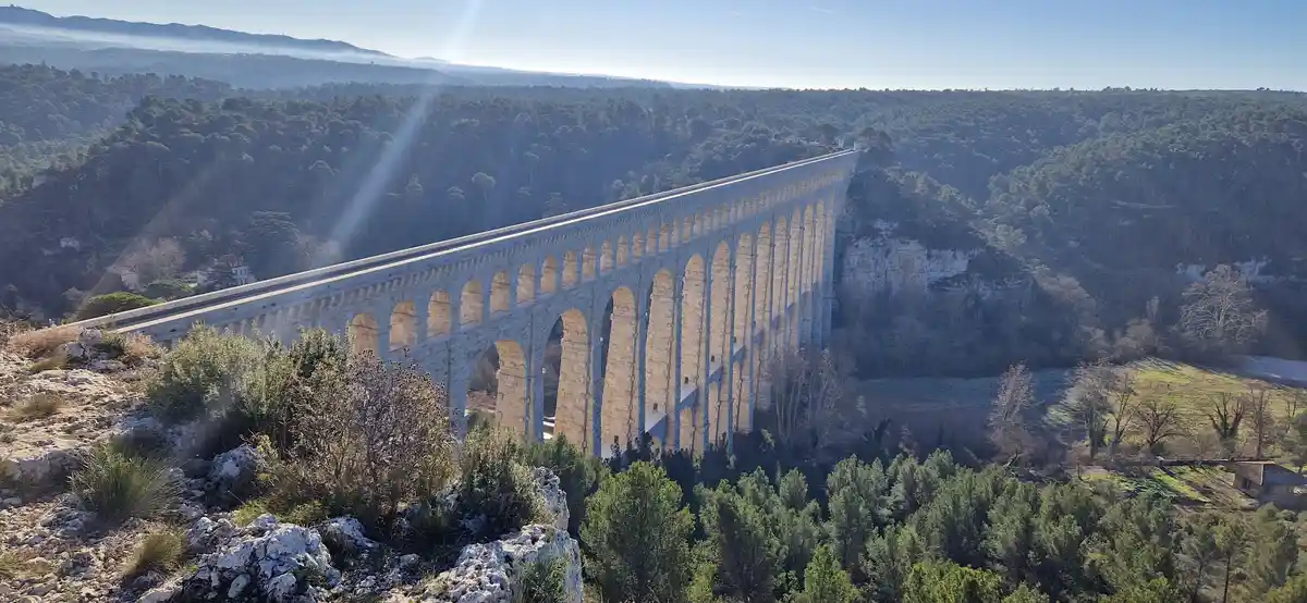 a wide-angle photograph shows the massive, arched stone structure of the Pont du Gard aqueduct spanning a deep, forested…