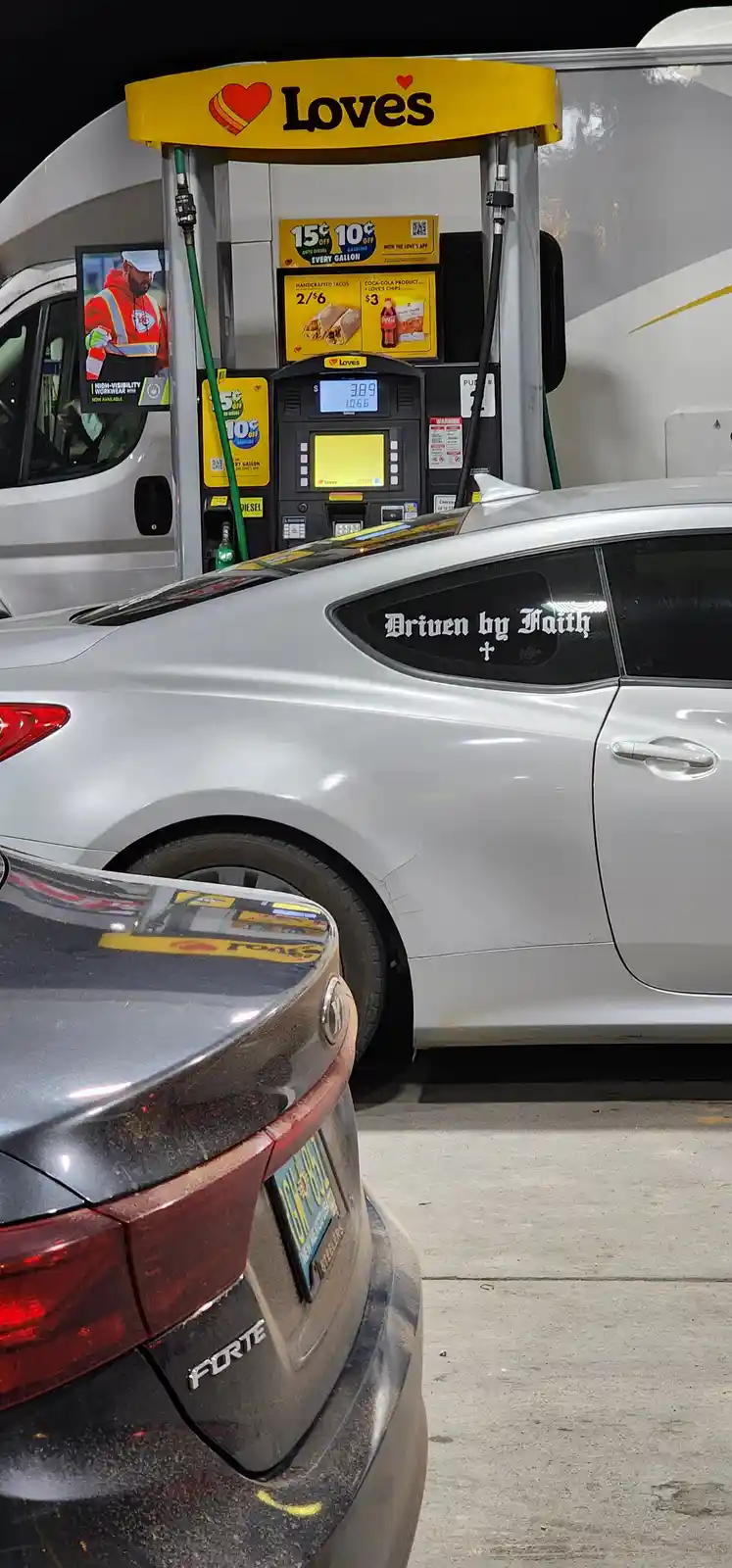 A silver car is parked at a Loves gas station at night, with a window decal reading 'Driven by Faith' and a small cross…