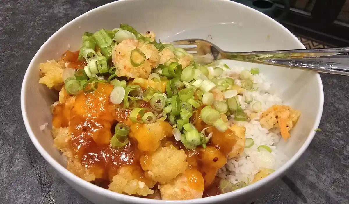 A white bowl contains fried shrimp covered in a glossy orange sauce and topped with chopped green onions, alongside a…