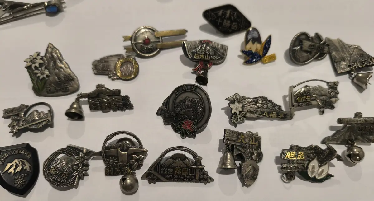 A collection of Japanese mountain climbing commemorative pins and badges, many shaped like mountains or featuring mountain…