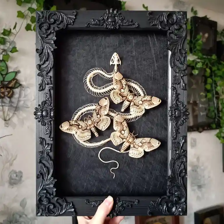 A hand holds a black ornate picture frame containing a 3D art piece of three golden moth sculptures intricately designed to…