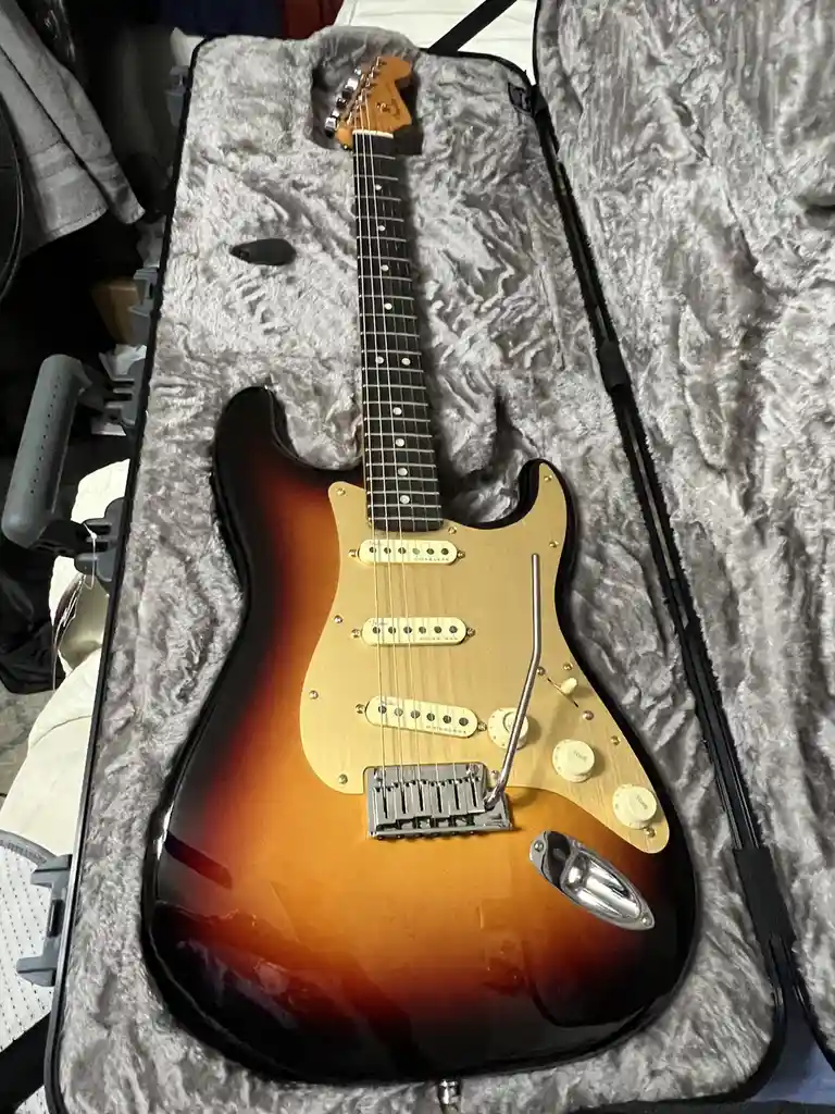 A sunburst-finish electric guitar with a maple neck and rosewood fretboard rests inside an open, grey plush-lined hard case…