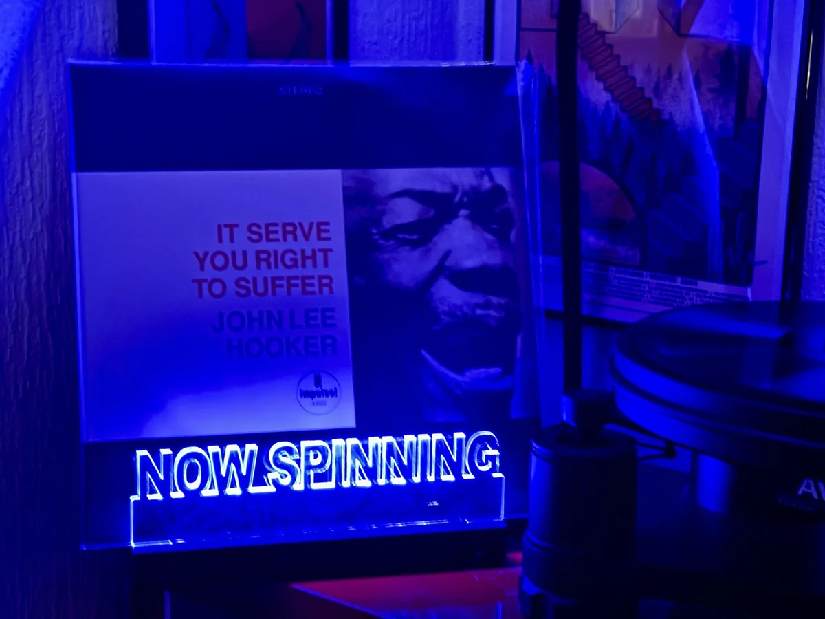 A vinyl record sleeve for John Lee Hooker's album 'It Serve You Right to Suffer' is displayed under blue lighting next to a…