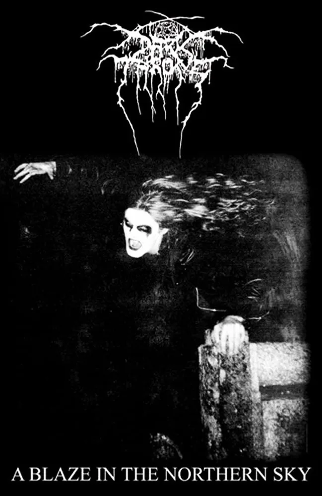 A black-and-white image shows a person with pale makeup, wild hair, and an intense expression, appearing to emerge from a…