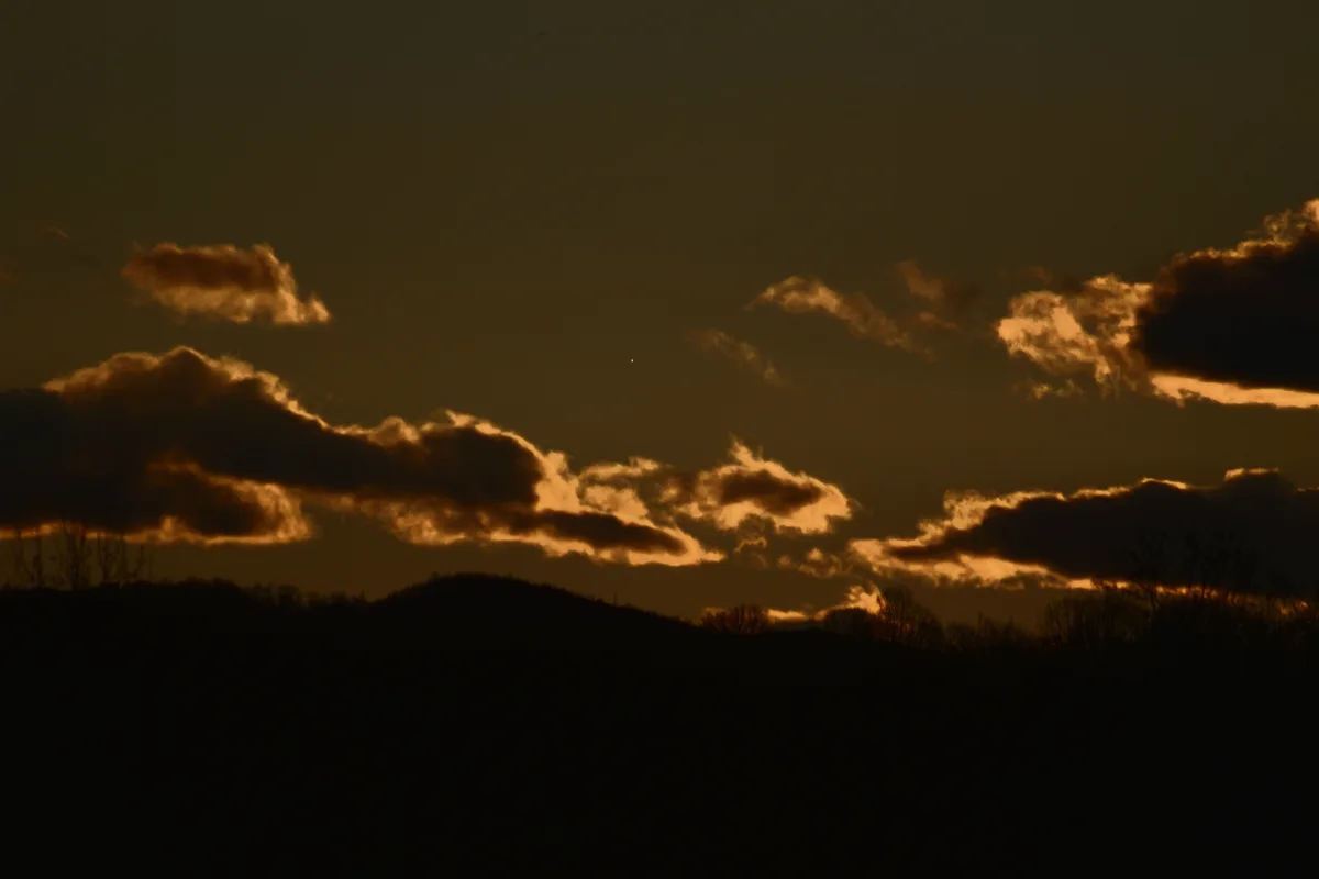 A serene sunset or sunrise photo showing dark silhouetted hills and bare trees against a sky filled with clouds edged in…