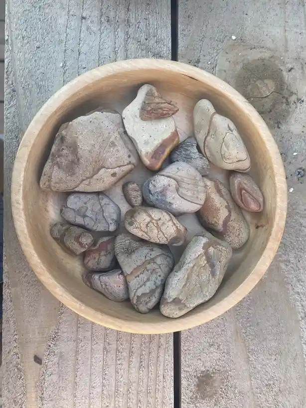 A top-down photo shows a light wooden bowl filled with various smooth, rounded stones of different sizes and earthy colors…