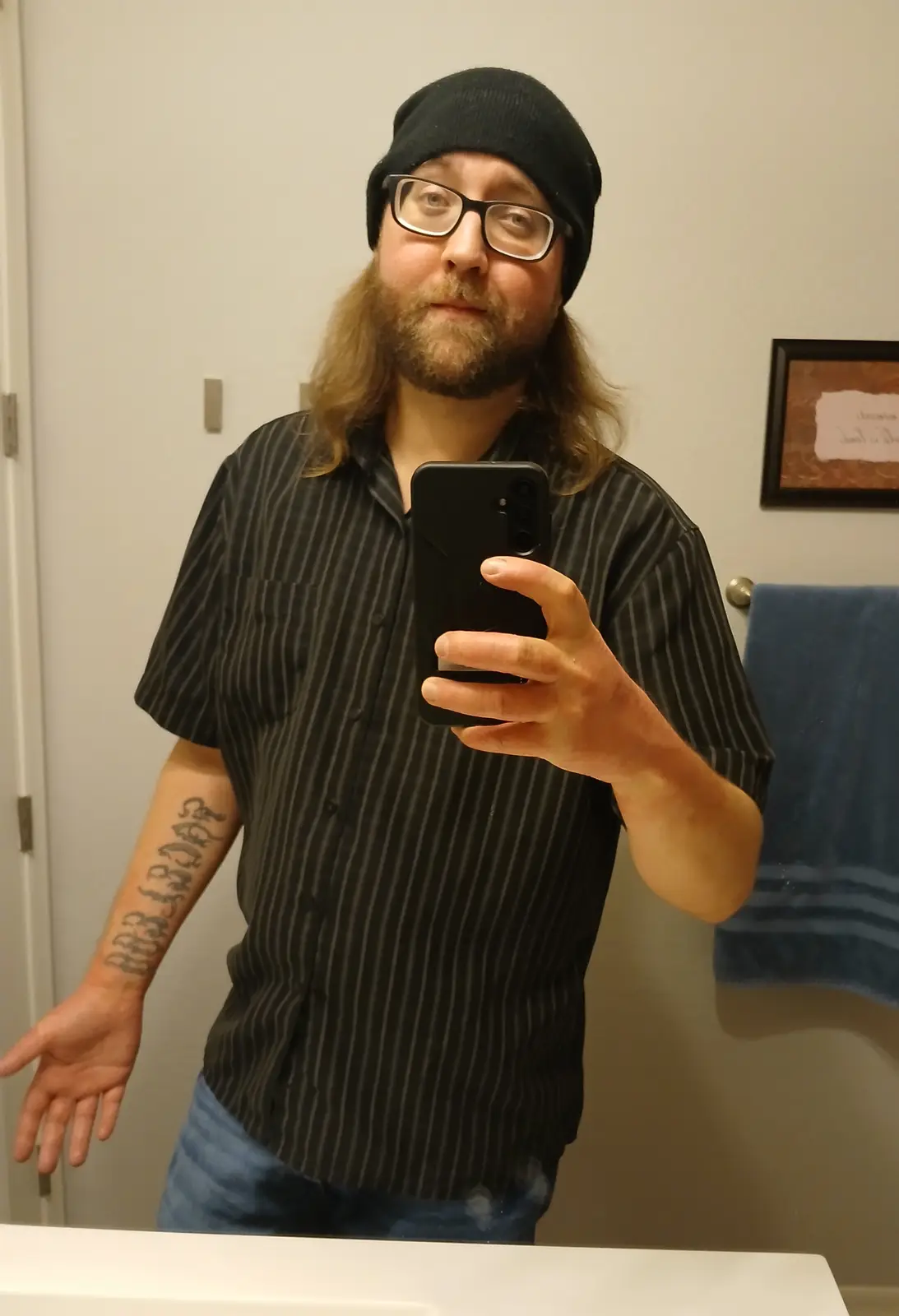 A man with a long beard, glasses, and a black beanie takes a mirror selfie in a bathroom while wearing a striped shirt and…