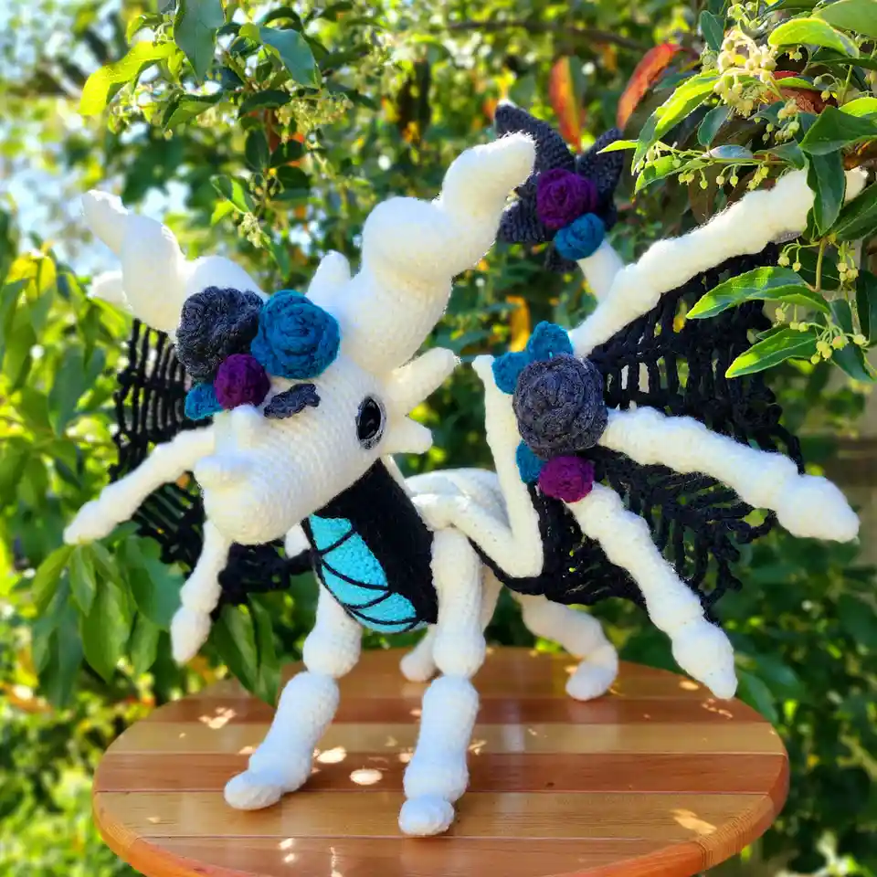 Two crocheted dragon figures, one with a mustache, stand on a wooden table outdoors against green foliage. Both have white…