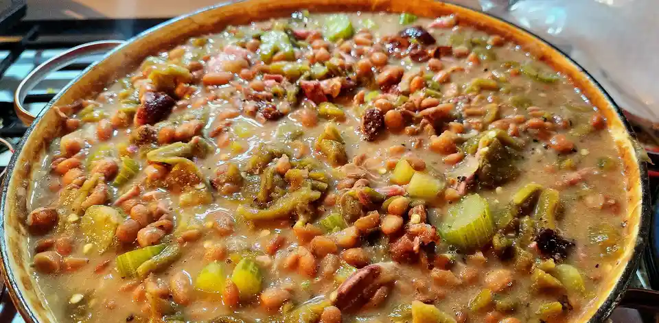 A close-up photo shows a large, round, shallow pan filled with a thick, creamy stew containing visible beans, chopped green…
