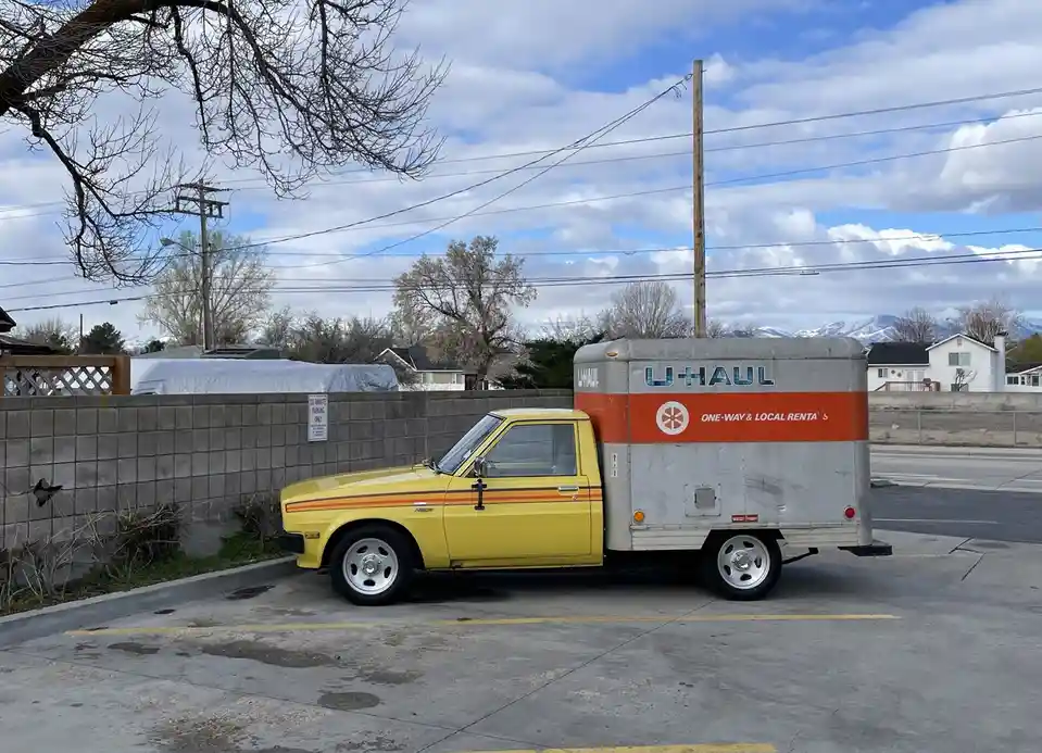 A vintage yellow pickup truck with an orange stripe is parked next to a U-Haul trailer in a lot. The trailer has the U-Haul…