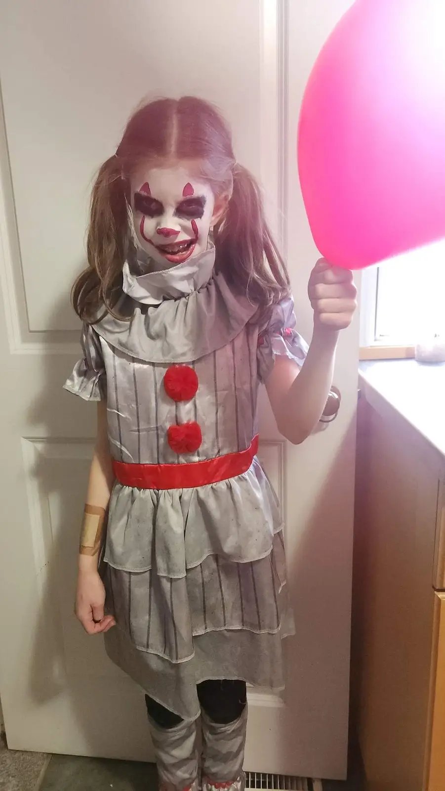 A young girl dressed as a scary clown with white face paint, red lips, and dark eye makeup, wearing a grey striped dress…