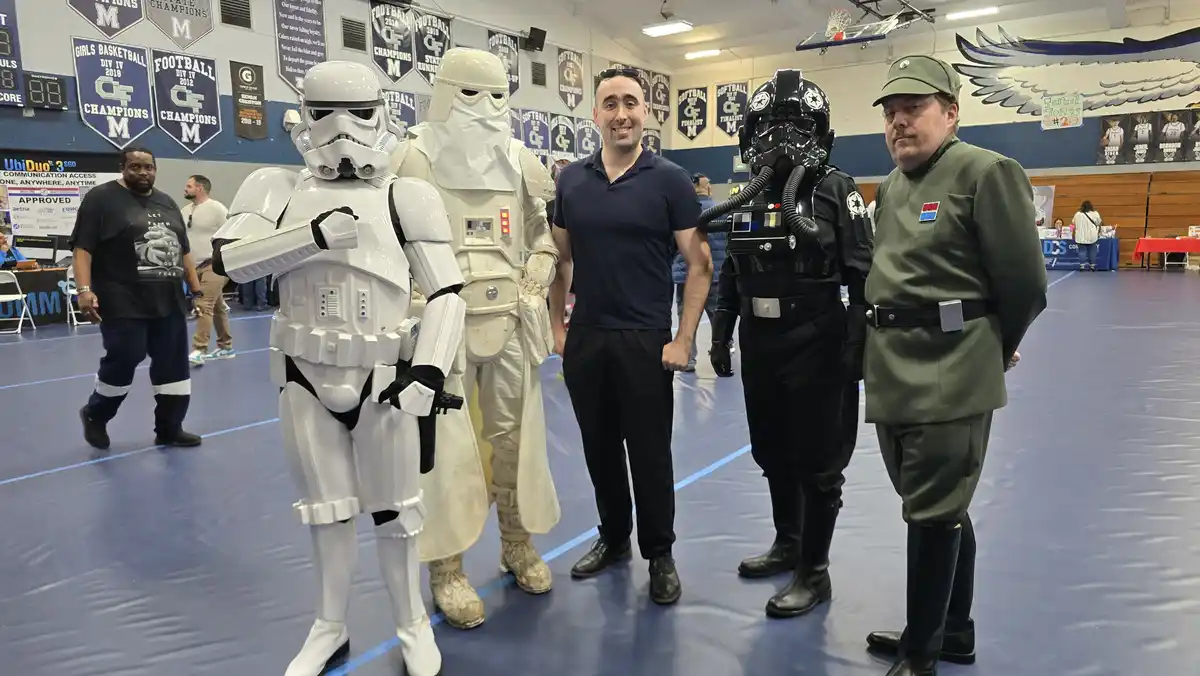 A man in a dark blue polo shirt stands smiling between two Star Wars cosplayers: a white-armored Stormtrooper and a…