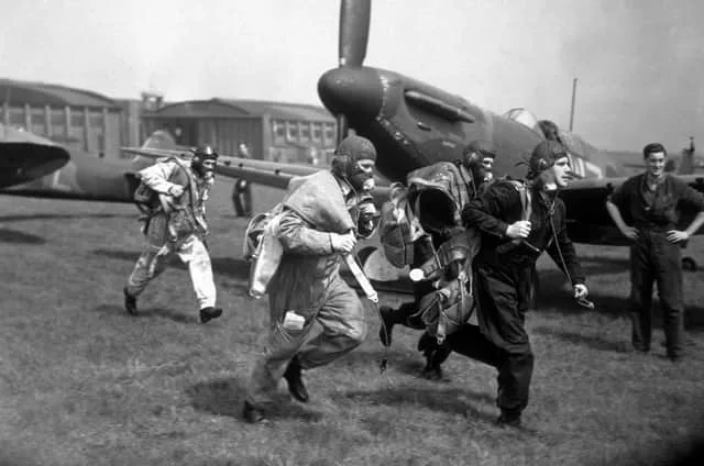 A black-and-white historical photo shows several WWII fighter pilots in full flight gear, running energetically toward a…