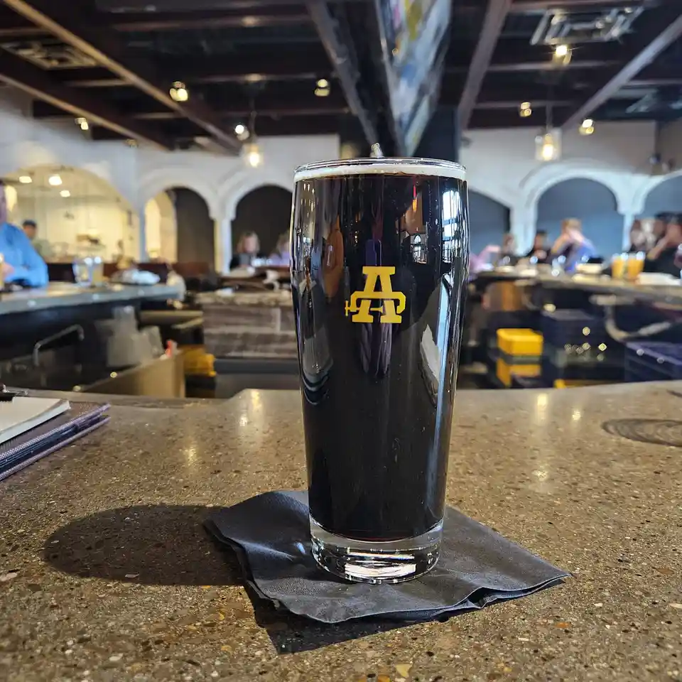 A tall glass of dark beer with a yellow 'A&T' logo sits on a black napkin on a speckled bar counter. The background shows a…