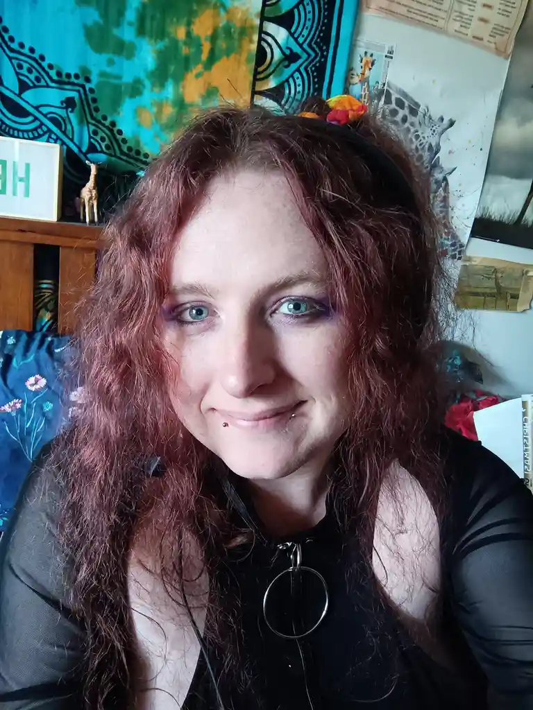 A person with long, curly reddish-brown hair and blue eyes smiles at the camera while wearing a black top with a circular…