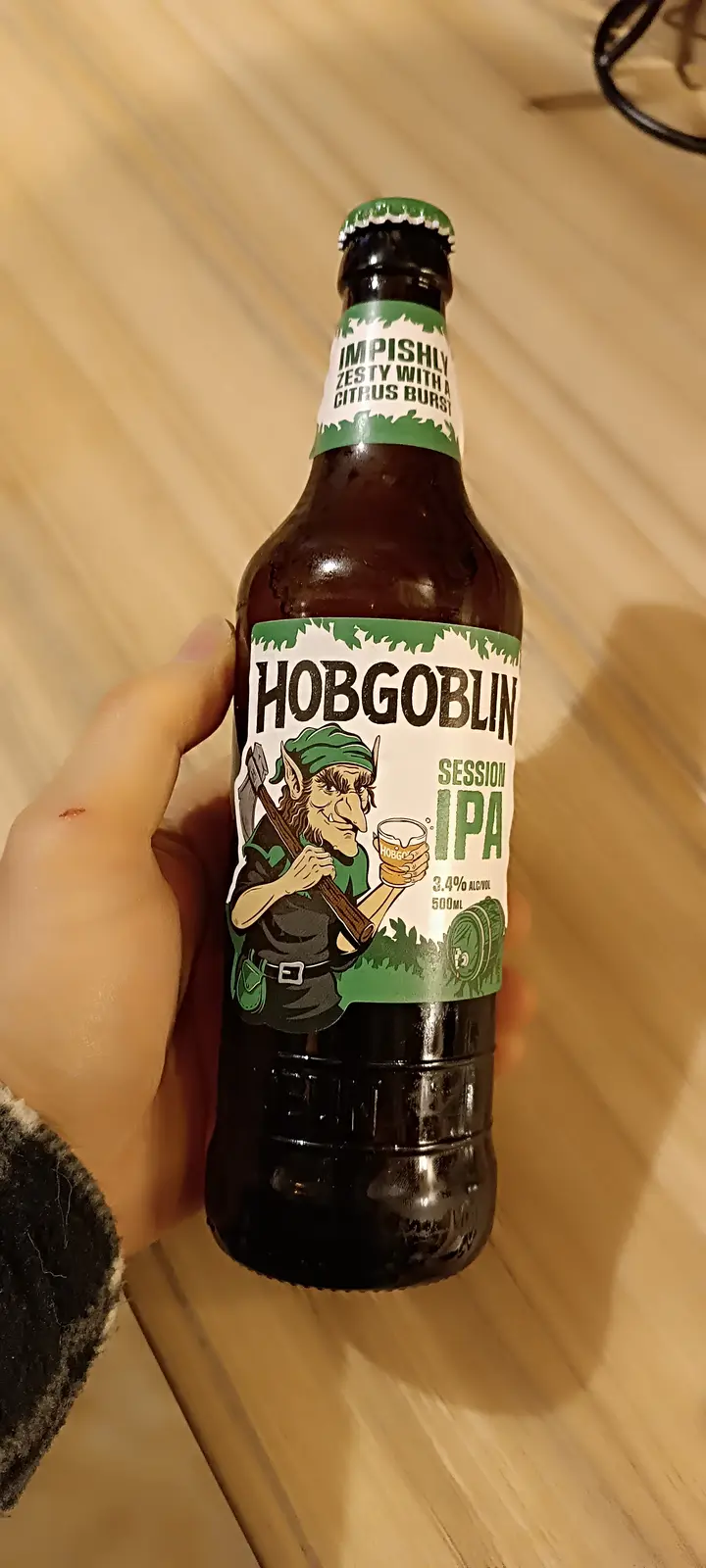 A hand holds a dark brown bottle of Hobgoblin Session IPA beer, featuring a label with a cartoonish green-hatted goblin…