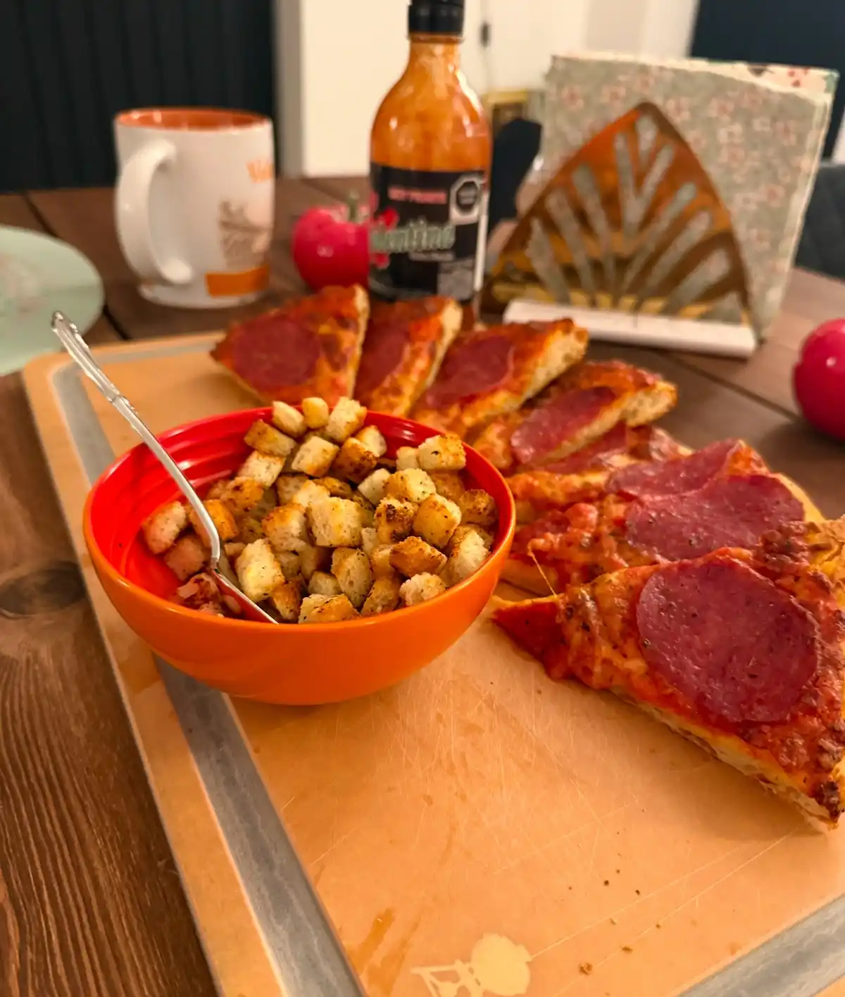 A close-up photo shows a wooden cutting board with several slices of pepperoni pizza and a red bowl of croutons. In the…