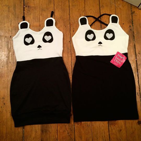 Two identical panda-themed dresses lie side by side on a wooden floor. Each dress has a white bodice with black ears, black…