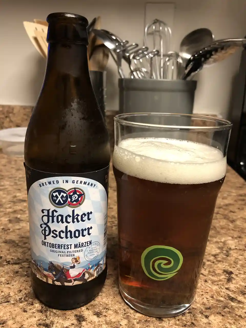 A brown glass bottle of Hacker Pschorr Oktoberfest Märzen beer sits next to a pint glass filled with the same beer, both…