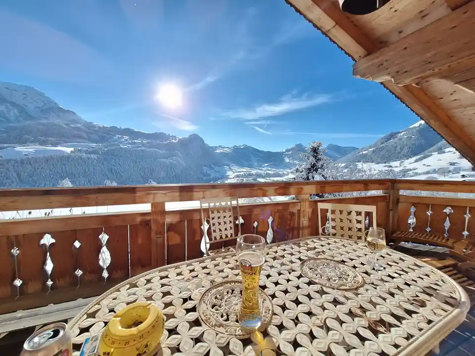A sunny winter scene from a wooden balcony overlooking snow-covered mountains, featuring a metal patio table with a beer…