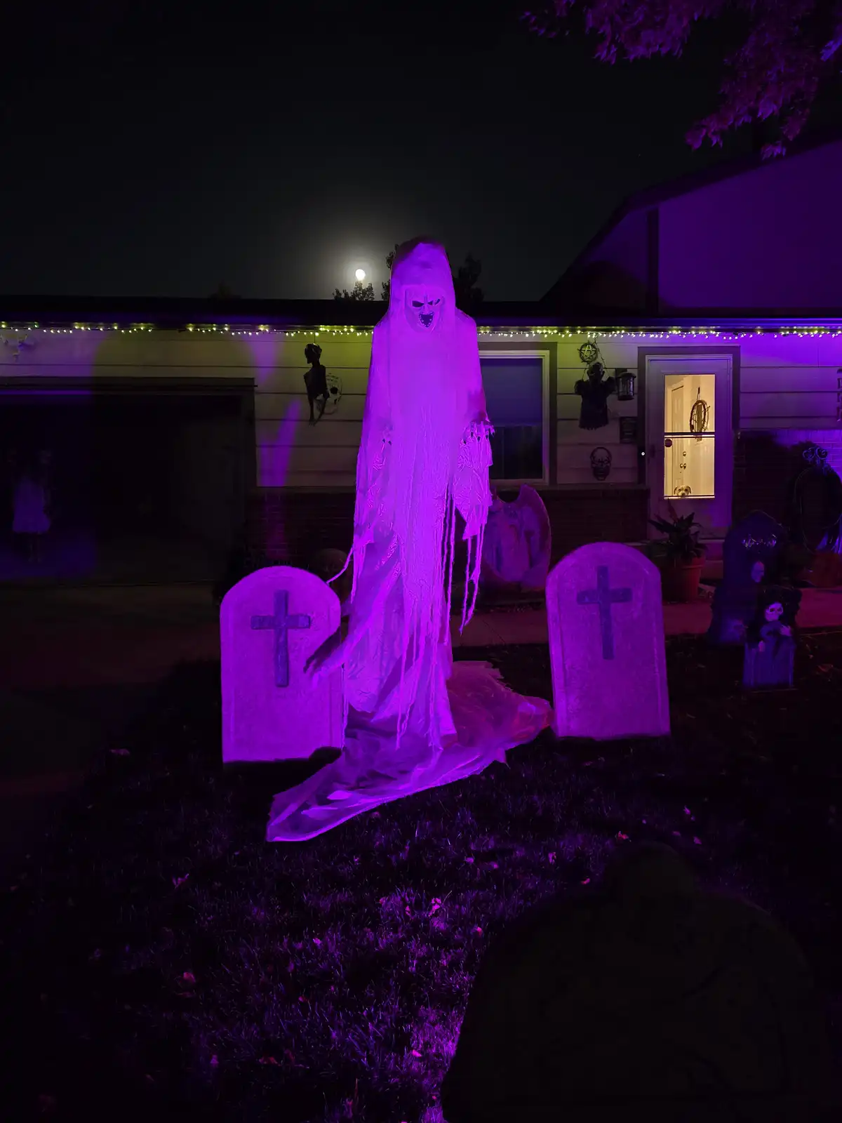 A large, glowing purple ghost decoration stands between two tombstones in a front yard decorated for Halloween, with a house…