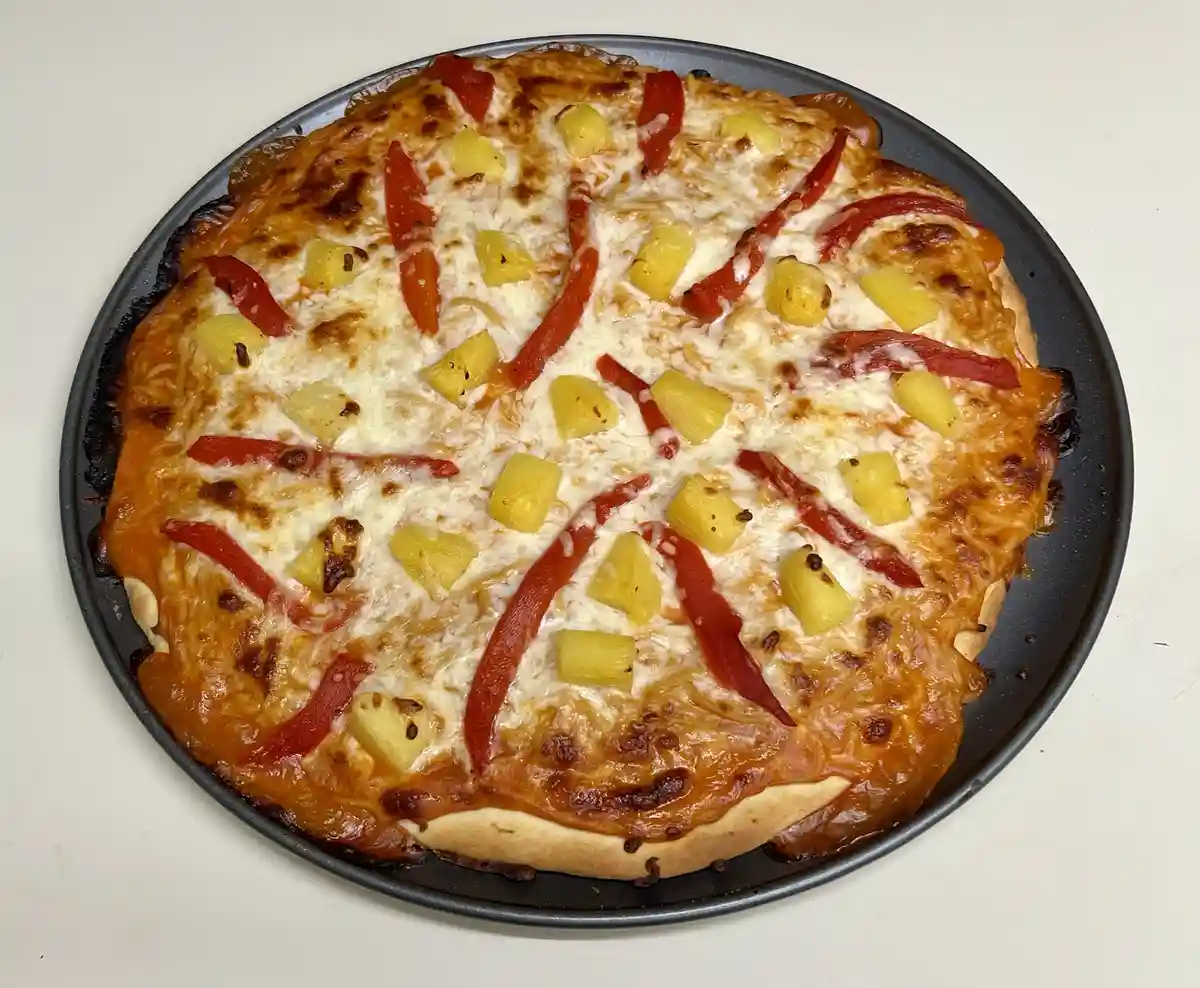 A freshly baked pizza on a black pan, topped with melted cheese, red bell pepper strips, and chunks of pineapple. The crust…