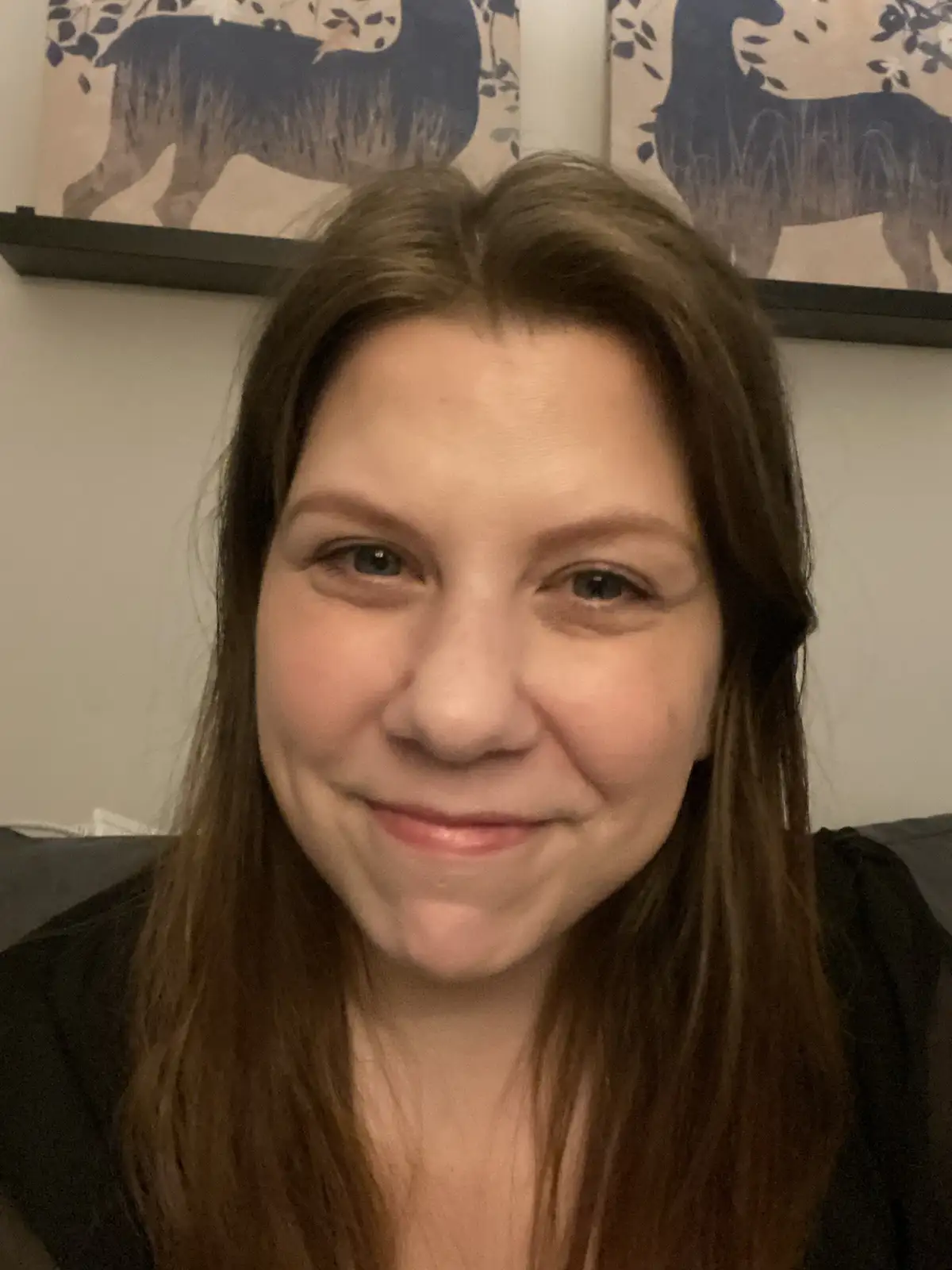 A close-up selfie of a woman with long brown hair and a subtle, slightly forced smile, looking directly at the camera…