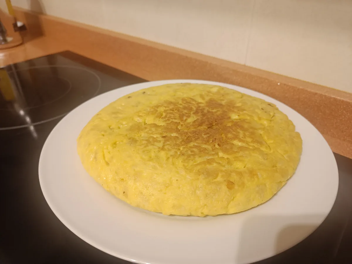A close-up photo shows a round, golden-yellow omelette resting on a white plate, placed on a dark kitchen countertop with a…