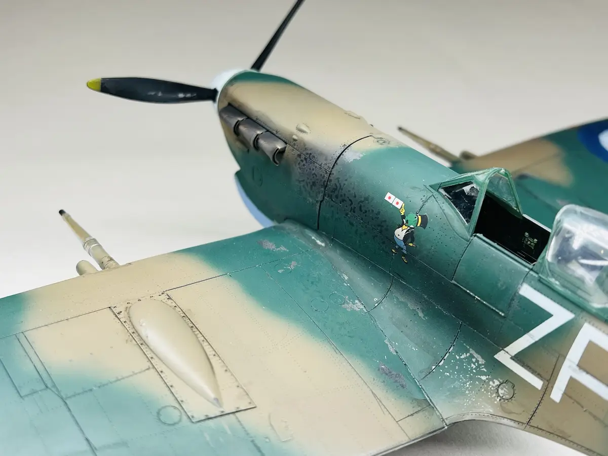 A close-up photo of a detailed model airplane with green and tan camouflage paint, showing its propeller, cockpit, and wing…