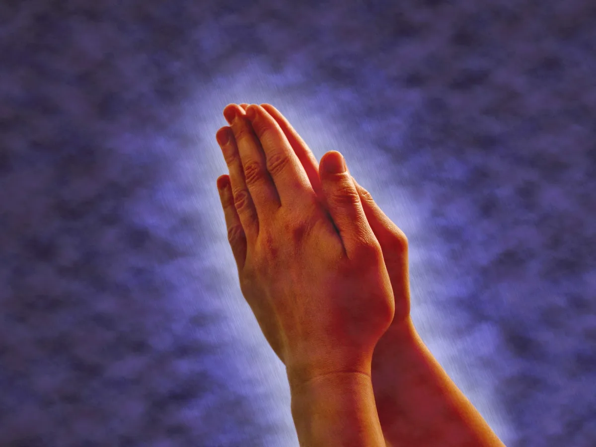 A pair of hands clasped together in a prayer-like gesture against a dark blue, cloudy sky background. The overlaid text…